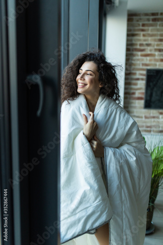 Beautiful woman standing in white blanket in light room, morning, relax time at home