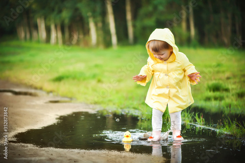 A baby girl in a yellow jacket plays with a rubber duckling in a puddle.
