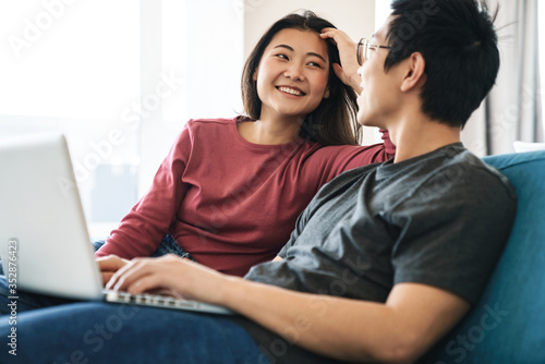 Photo of asian couple laughing and using laptop while sitting on sofa