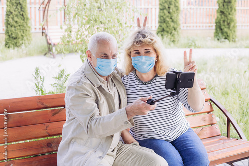 Senior couple in the park wearing medical mask to protect from coronavirus and making selfie in spring or summer day, coronavirus quarantine