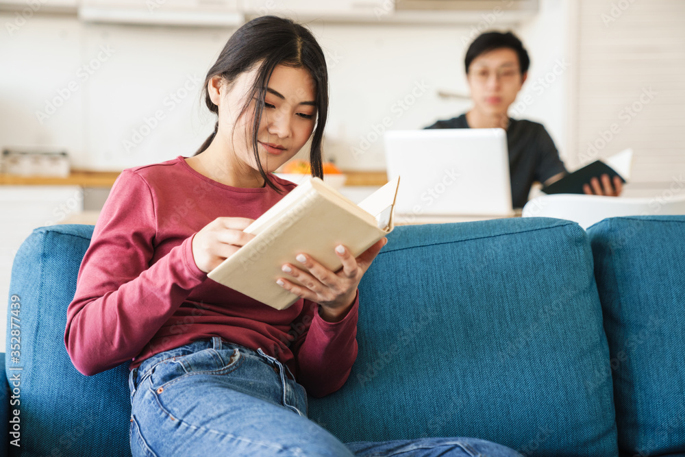 Photo of young concentrated asian couple reading books and using laptop Photo of young concentrated asian couple reading books and using laptop