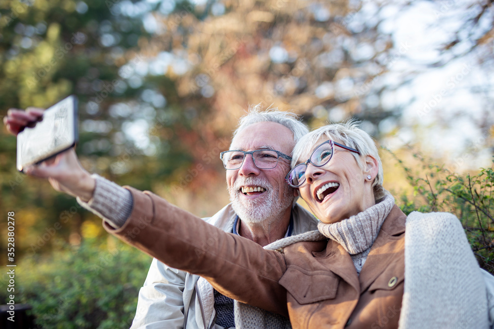 Fototapeta premium MAture couple taking selfie by mobile phone while sitting on bench in public park