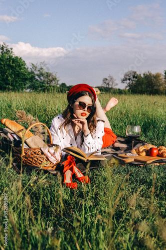 Stylish modern summer time picnic in nature. Beautiful girl in red hat and black sunglasses in sunny day read book and enjoys the moment