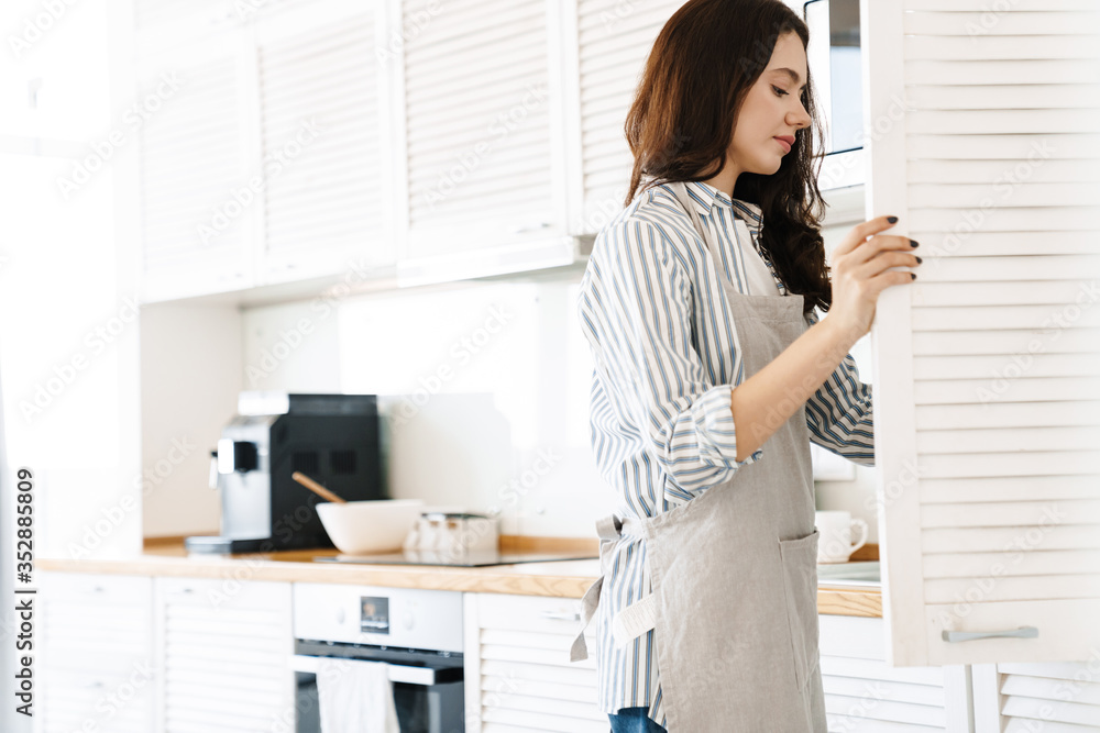 Image of focused young woman opening cupboard while cooking pie