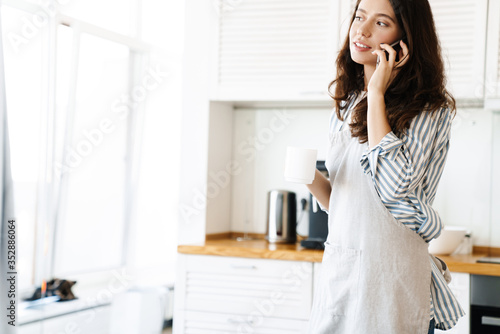 Image of young beautiful woman talking on cellphone while cooking pie