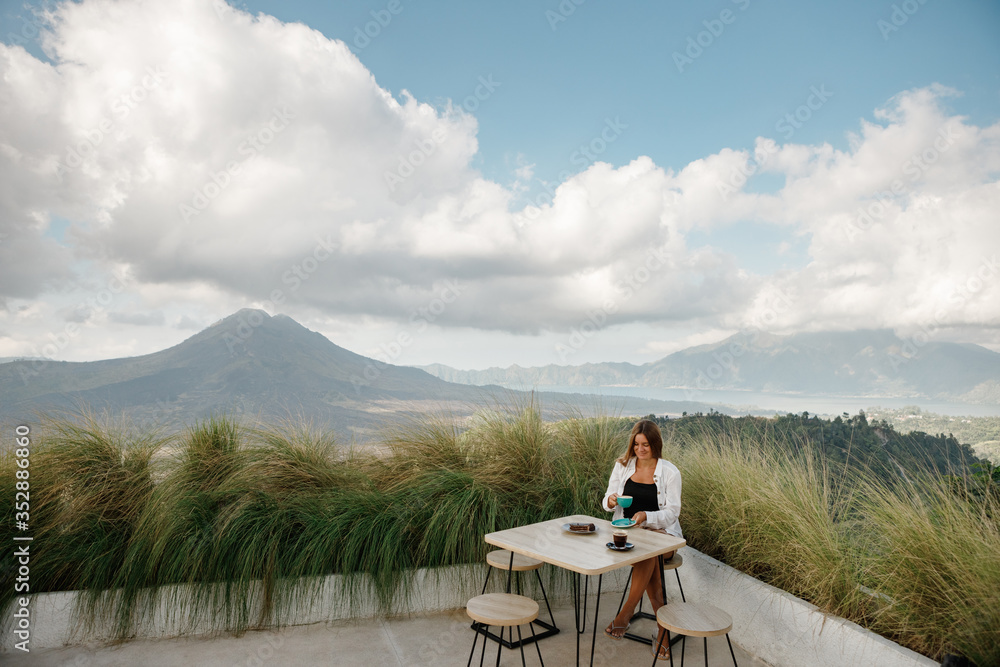 Coronavirus is over. Woman drinking warm tea in the rustic wooden outdoor cafe with mountain view. Young girl traveler sits on a wooden veranda, enjoys the mountain scenery, drink coffee