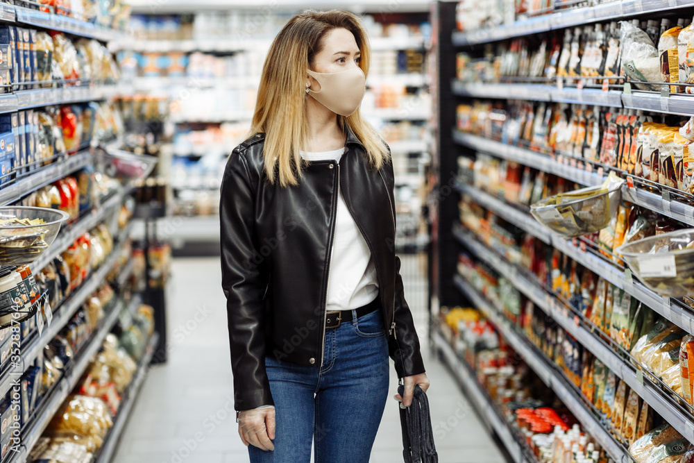 Pretty young woman wearing face mask and rubber gloves in suppermarket departmentstore. Girl choosing, looking grocery things to buy at shelf during coronavirus crisis or covid19 outbreak.
