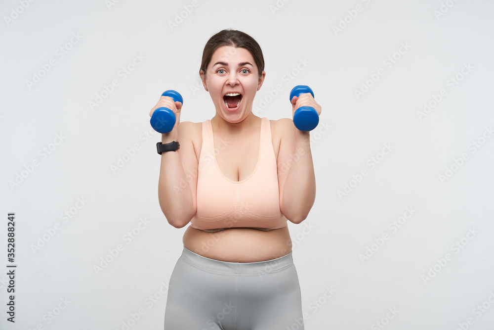 Indoor photo of young brunette plump lady with chubby face lifting dumbbells and looking excitedly at camera while posing over white background. Bodycare concept Indoor photo of young brunette plump lady with chubby face lifting dumbbells and looking excitedly at camera while posing over white background. Bodycare concept