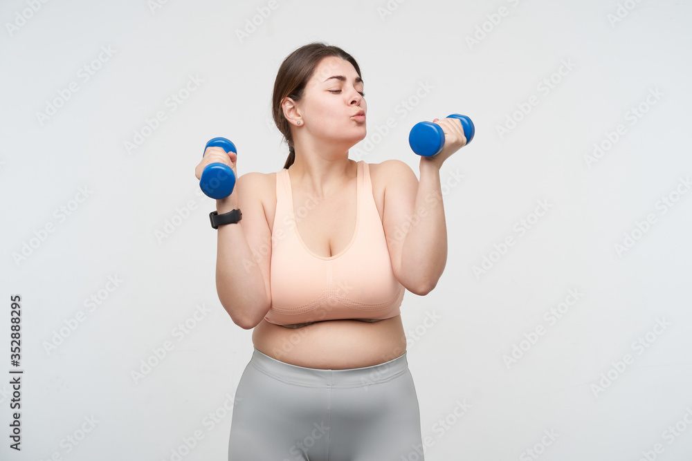 Positive young oversized female dressed in sports bra and leggins keeping blue dumbbells in raised hands and forming lips in kiss while looking at it, isolated over white background