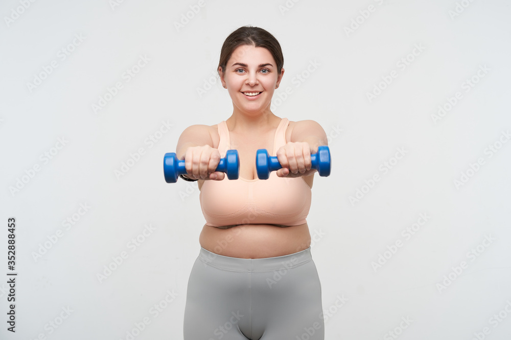 Studio shot of young pleased oversized female making static exercises with weight and smiling gladly at camera, working out her body while posing over white background Studio shot of young pleased oversized female making static exercises with weight and smiling gladly at camera, working out her body while posing over white background