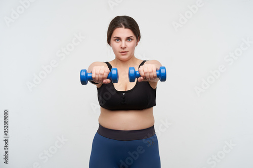 Concentrated young brunette chubby lady in sports bra and leggins keeping hands in front of herself while making exercises for hands, posing over white background