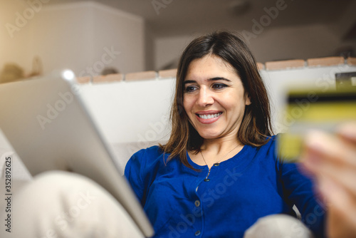 Young caucasian happy woman sitting on the sofa doing online shopping with tablet in her free time. Internet easy payment by credit card. Technology and home banking concept.