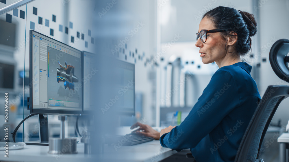 Industrial Female Engineer Working on a Personal Computer, Two Monitor Screens Show CAD Software with 3D Prototype of New Hybrid Hydrogen Engine and Charts. Modern Factory with High-Tech Machinery