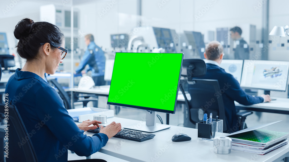 Over the Shoulder: Female Chief Engineer Working on Personal Computer, Display is Green Mock-up Screen or Chroma Key. Industrial Factory with High-Tech Machinery and Professional Workers Over the Shoulder: Female Chief Engineer Working on Personal Computer, Display is Green Mock-up Screen or Chroma Key. Industrial Factory with High-Tech Machinery and Professional Workers