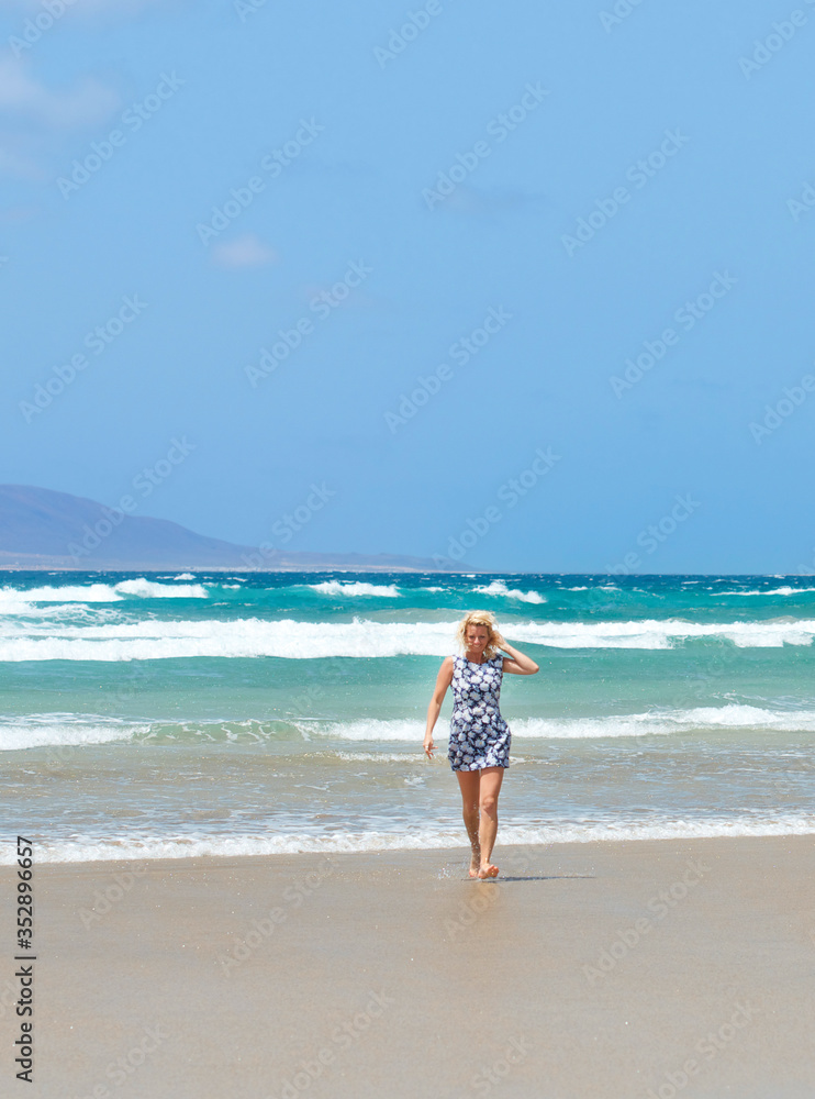 Blonde caucasian white woman in blue short dress walking and enjoying sunny day In Famara beach in the northwest of the island of Lanzarote,one of the most popular places to surf in the Canary Islands Blonde caucasian white woman in blue short dress walking and enjoying sunny day In Famara beach in the northwest of the island of Lanzarote,one of the most popular places to surf in the Canary Islands