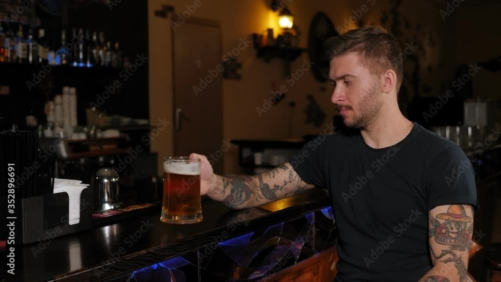 A bearded young man with tattoos drinking beer at bar or pub. People, drinks, alcohol and leisure concept. A bearded young man with tattoos drinking beer at bar or pub. People, drinks, alcohol and leisure concept.