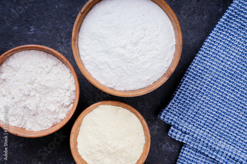 Rice flour, corn flour and whole grain flour with blue napkin in wooden bowls on dark background. Baking ingredients. Flat lay top view, studio shot