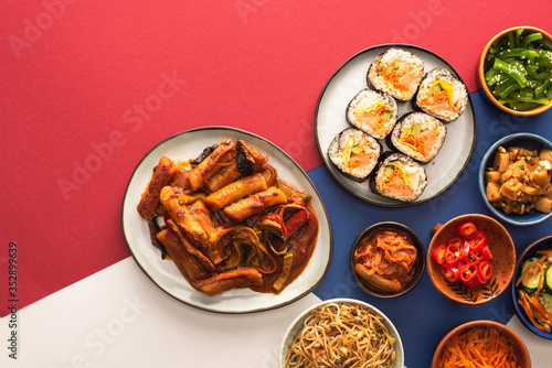top view of korean topokki near bowls with spicy side dishes on blue, crimson and white
