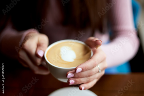 Close-up picture of female hands with pink manicure, holding white coffee mug with cappuccino foamy hot drink. Sunday leisure time. Food and drink establishment.