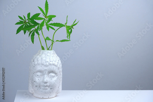 White Buddha's head with green marijuana leaves on a gray background with copy space