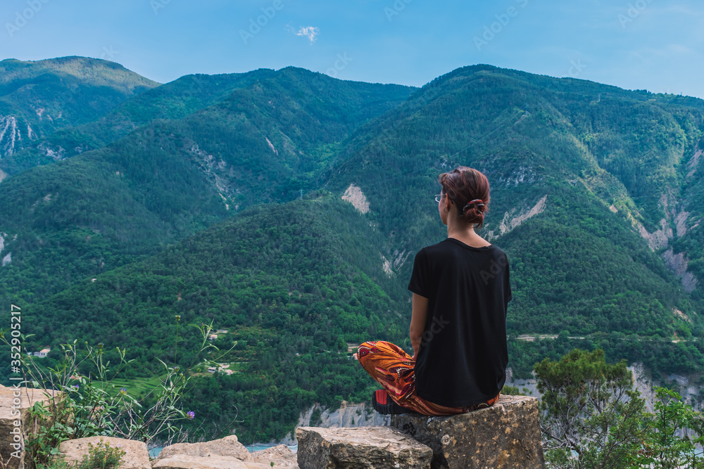 A full body shot of an unrecognizable young Caucasian redhead hiker sitting on a rock enjoying the view of the valley of Var in the French Alps mountains
