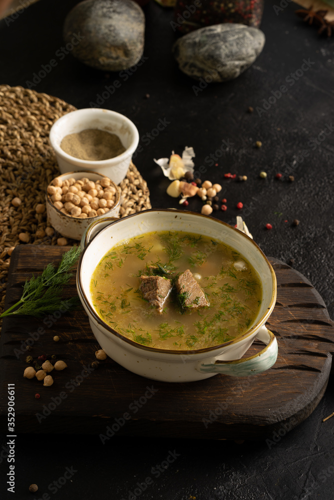 Vegetable soup with beef, potatoes and dill garnished with garlic, chickpeas, black pepper and bay leaf. A bowl of soup and ingredients on a black table. Vegetable soup with beef, potatoes and dill garnished with garlic, chickpeas, black pepper and bay leaf. A bowl of soup and ingredients on a black table.