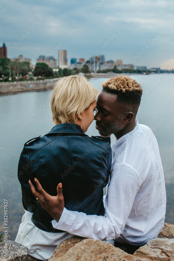 Interracial couple sits on rocks and hugs against background of river and city. Interracial couple sits on rocks and hugs against background of river and city.