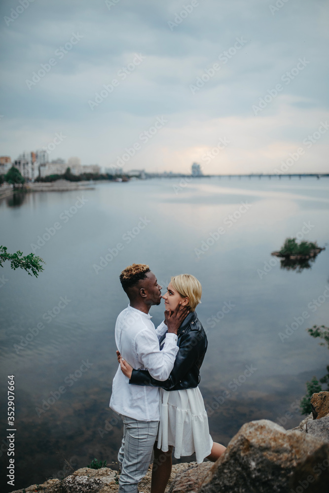 Interracial couple stands and hugs against background of river and city. Interracial couple stands and hugs against background of river and city.