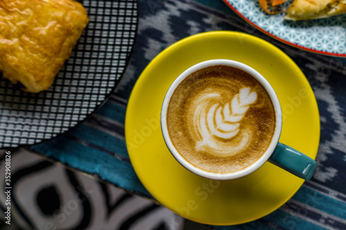 A cup of latte is served with colorful ceramic cups  side by side with food and cutlery. Everything is photographed on the table with traditional woven cloth ornaments ..