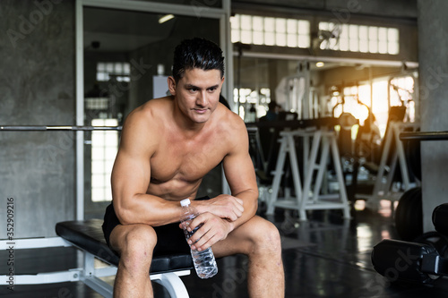 Handsome muscular man sitting and drinking water after his workout in the fitness gym