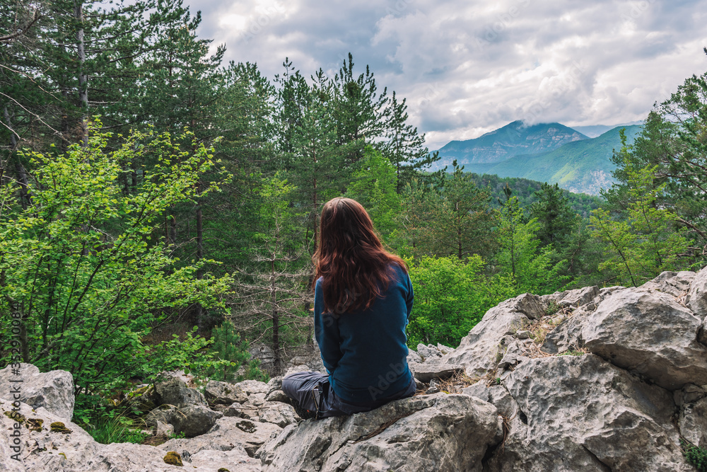 Naklejka premium A full body shot of an unrecognizable young Caucasian female hiker sitting on a rock in the French Alps mountains on a cloudy summer day