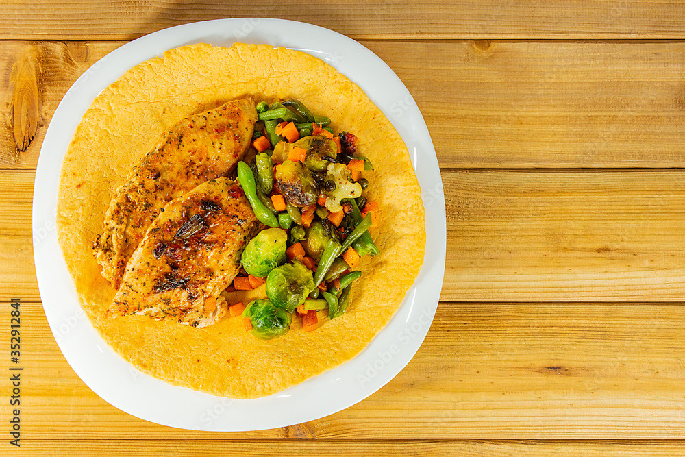 Grilled chicken breast and a mixture of vegetables on a corn tortilla in a white plate with copy space on a wooden background. Close-up Grilled chicken breast and a mixture of vegetables on a corn tortilla in a white plate with copy space on a wooden background. Close-up
