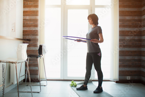 Adult fit slim woman has workout at home. Senior concentrated senior adult holding hula hoop around waist and ready to exercise. Take care about good shape and figure.