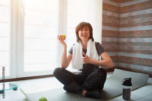Fototapeta Naklejka Na Ścianę i Meble -  Adult fit slim woman has workout at home. Sit alone on yoga mat with apple in hand. Healthy snack after exercising. Well-built strong woman on picture.