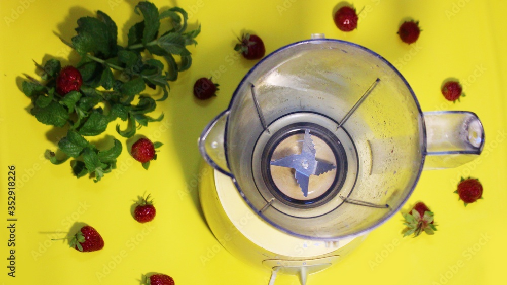 Milk and strawberry smoothie is mixed in a mixer on a yellow background with mint. Strawberries and milk are mixed in a blender on the table. View from above. Summer fresh cocktail mixed in a blender