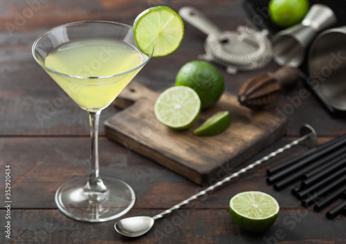 Gimlet Kamikaze cocktail in martini glass with lime slice and ice on wooden board with fresh limes and strainer with shaker.