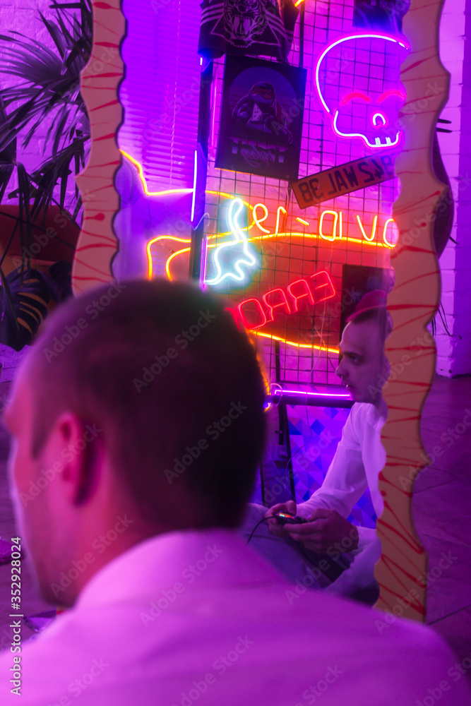 Fototapeta Portrait of the Young Handsome gamer. Reflection of the man on the mirror. Neon Colored Room. Old 90's retro style. Reflect of neon words near male. Selective focus.