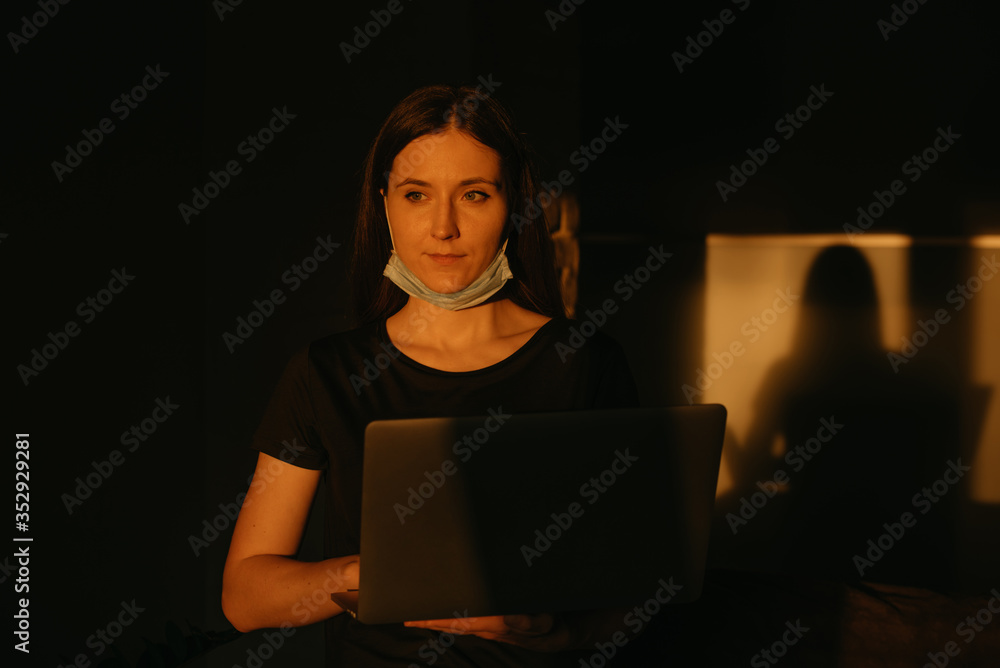 A woman in the removed medical face mask working remotely on her laptop during the quarantine to avoid the spread coronavirus. A lady working from home in the evening during the pandemic of COVID-19. A woman in the removed medical face mask working remotely on her laptop during the quarantine to avoid the spread coronavirus. A lady working from home in the evening during the pandemic of COVID-19.