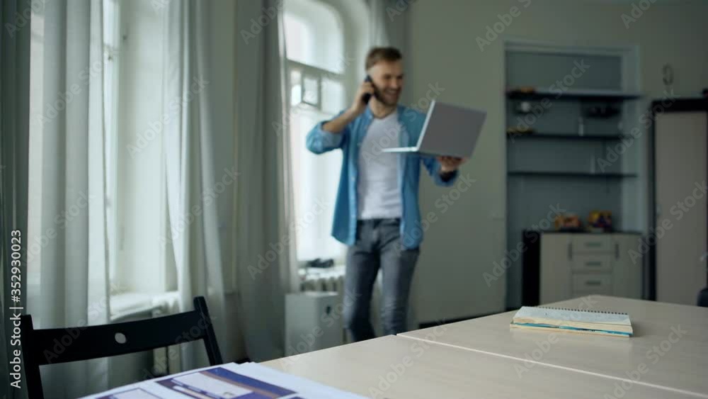 Smiling excited young man with laptop computer sitting down at desk while having business call on cell phone. Businessman finishing talk, closing pc and day dreaming at home Smiling excited young man with laptop computer sitting down at desk while having business call on cell phone. Businessman finishing talk, closing pc and day dreaming at home