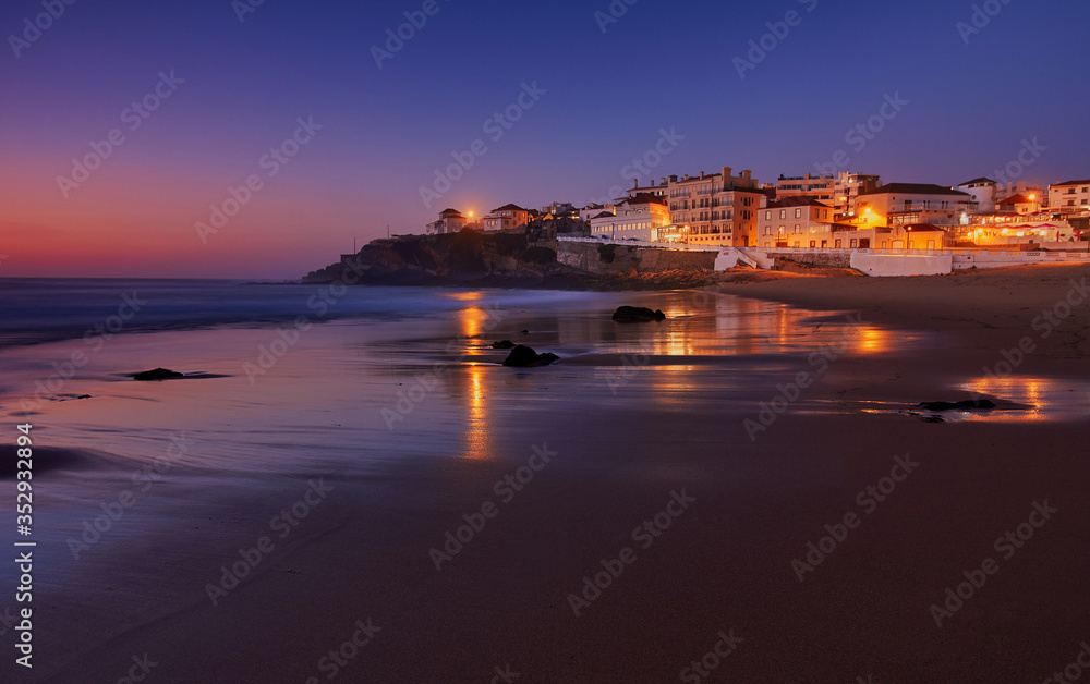 Amazing landscape of the Atlantic ocean coast at dusk. Night view on the village in lights, reflecting on a sandy beach. Long exposure image. Beach of Praia das Macas. Sintra. Portugal.