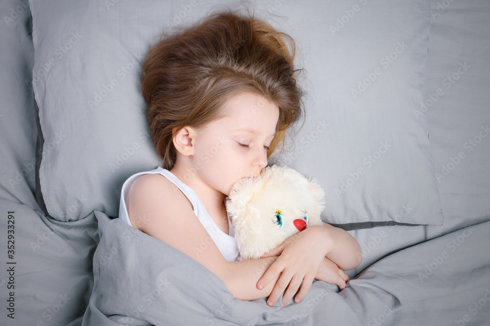 A little girl with brown hair is sleeping on her side with a stuffed toy in the bed under the blanket. Solid gray bed linen top view close-up.