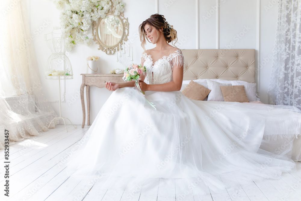 A gorgeous bride in a beautiful dress is sitting on a bed in a bright room and holding a bouquet of flowers . Wedding. Wedding bouquet. morning of the bride. A happy bride is waiting for the groom. A gorgeous bride in a beautiful dress is sitting on a bed in a bright room and holding a bouquet of flowers . Wedding. Wedding bouquet. morning of the bride. A happy bride is waiting for the groom.