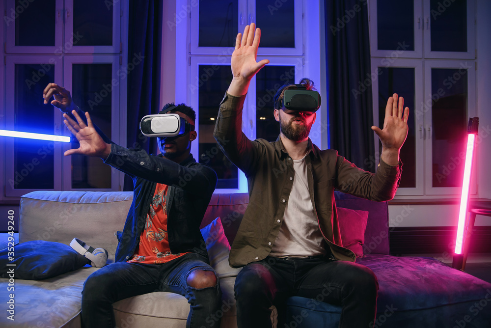 Fototapeta premium Portrait of positive likable satisifed multiracial male mates which sitting together on the couch in somebody's room and competing between each other in video game using special 3d goggles