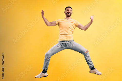 That's cool ! Young man wearing a yellow t-shirt and light jeans jumping against a yellow wall background and holding his thumbs up.