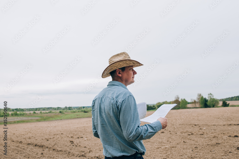 Man farmer agronomist in jeans and shirt stands back in field after haymaking, with tablet looking into the distance. Rural business, agricultural industry, freedom after work, concept