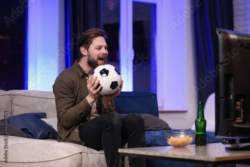 Good-looking modern positive 30-aged guy with well-groomed beard savoring chipps during emotional review of favourite football team's game on tv at home photo