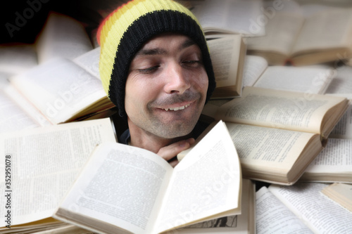 A young man in a hat reads a lot of books at the same time. Only the head sticks out of a huge number of open books. Selective focus.
