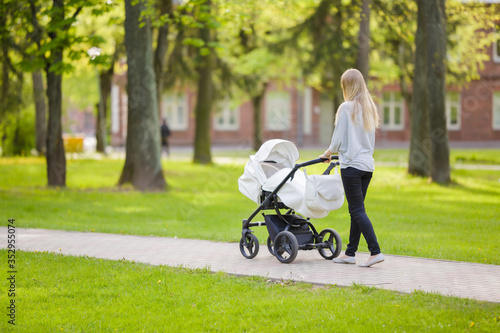 Картината върху платно One young mother pushing white baby stroller and slowly walking at town green park in warm, sunny spring day