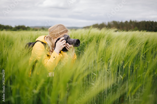 Charming blonde photographer in yellow jacket taking pictures of green wheat field. Mature lady using professional digital camera for creating amazing photos on fresh air.
