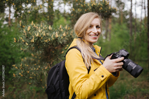 Attractive woman in yellow jacket with backpack standing at green forest, smiling and looking at camera. Female photographer with blond hair holding digital camera outdoors. © Kuz Production